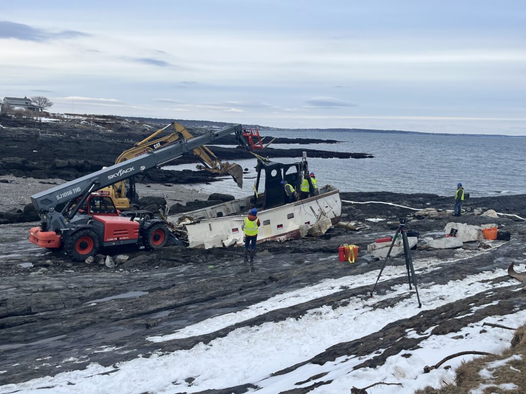 Upright hull with most accessories removed, and two excavators working on removing the next piece.